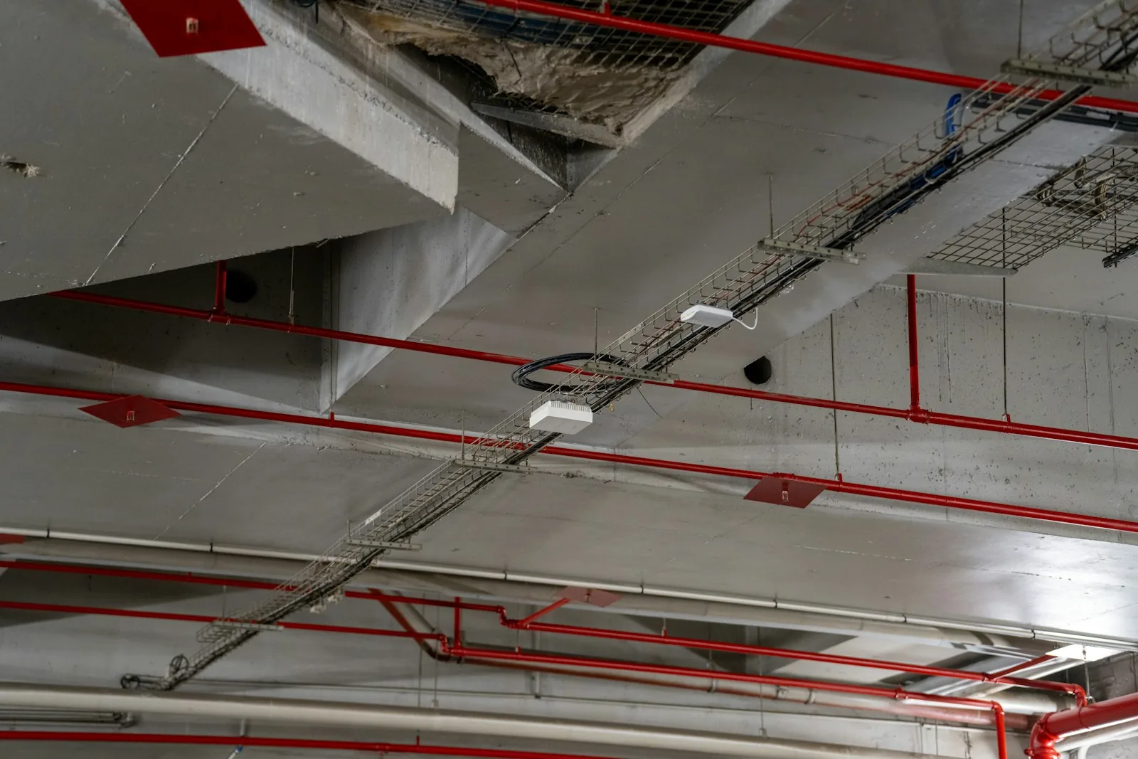 Exposed building services ceiling with concrete beams, red pipework and cable trays