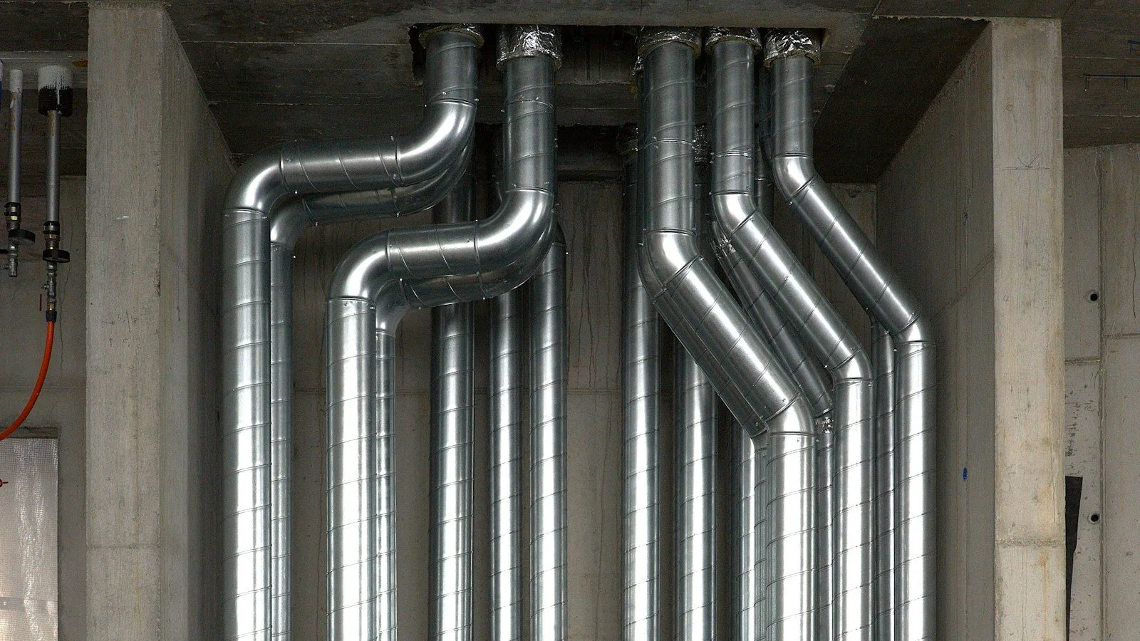 Bundled galvanised duct risers and pipework running up a bare concrete mechanical shaft